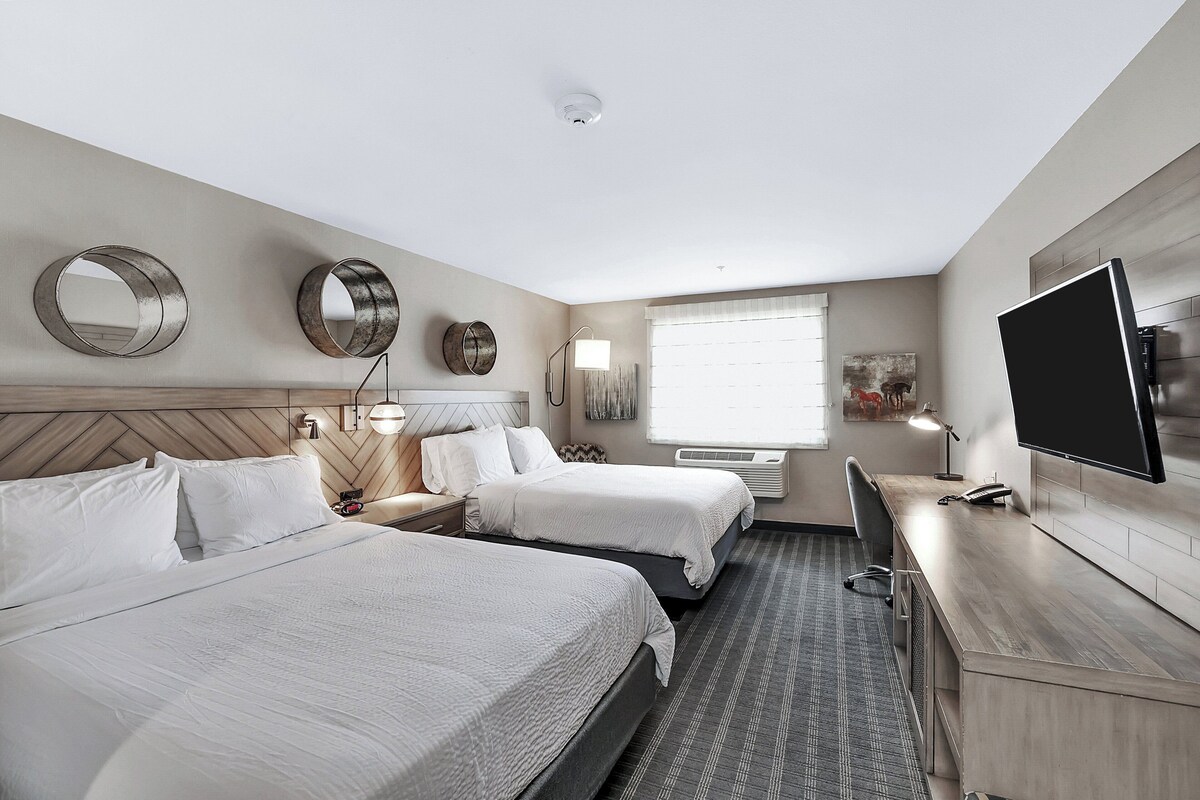 A hotel room with two beds, a desk, a flat-screen TV, and a large window with blinds.