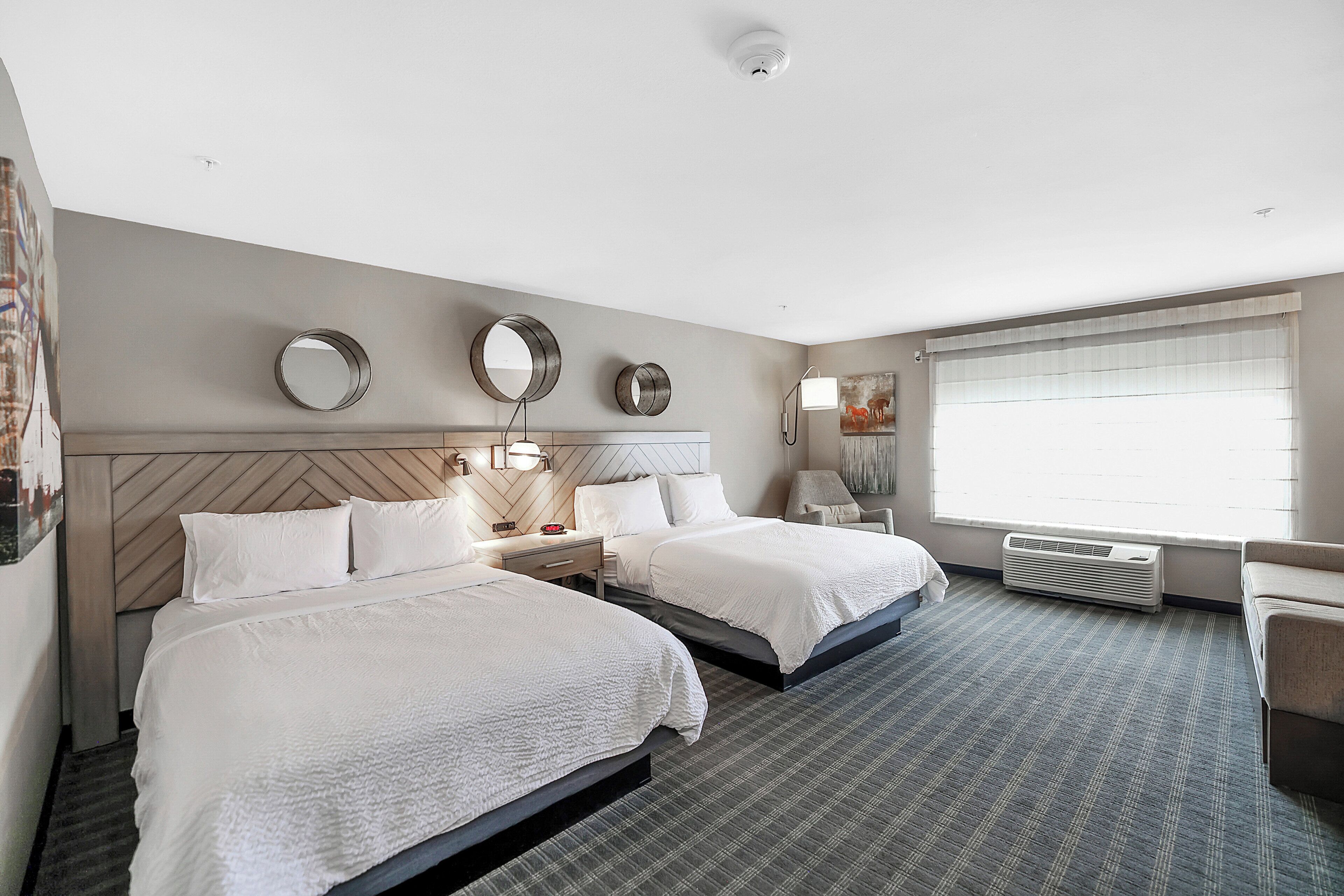 A hotel room with two beds, a large window with blinds, a wall-mounted air conditioner, and a headboard with a geometric pattern.