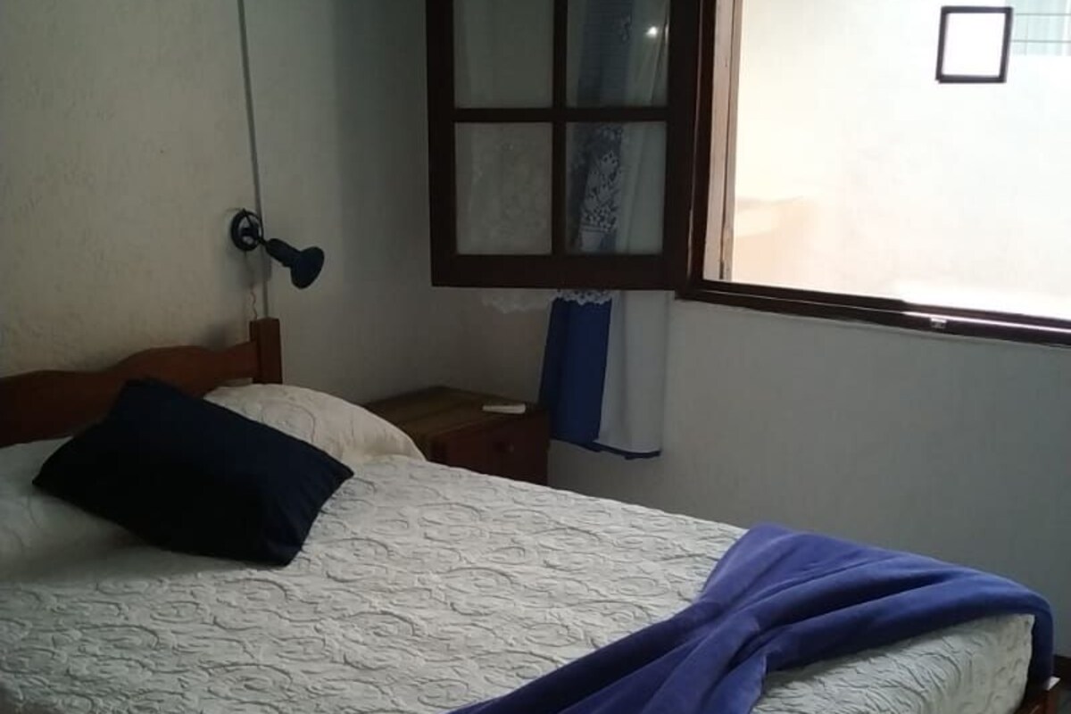 A bedroom with a bed, a window with curtains, a wall-mounted air conditioner, and a power outlet.