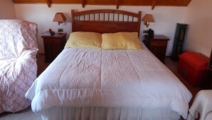 Un dormitorio con techo de madera, cama con ropa de cama blanca y amarilla, una silla con estampado floral y una mesita de noche con lámpara.