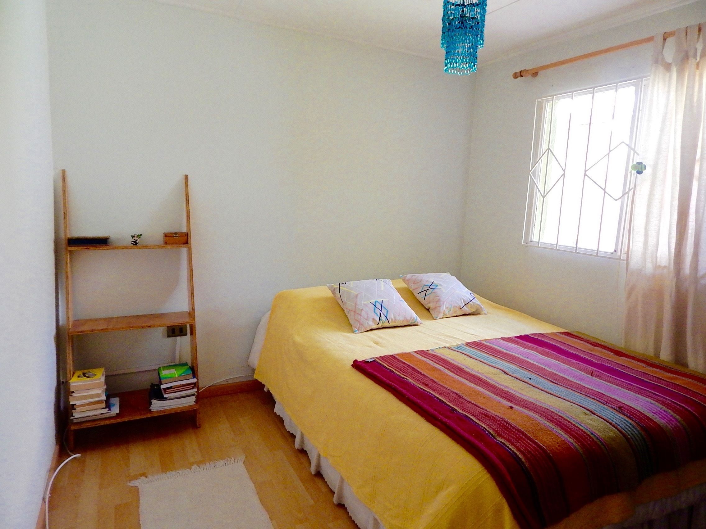 A small, bright room with a bed, a wooden shelf with books, and a window with curtains.