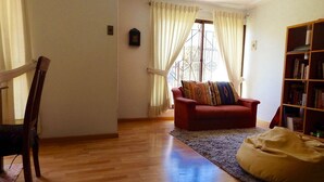 Una sala de estar con piso de madera, un sofá rojo, un sillón de peluche amarillo, una estantería y un ventanal con cortinas.
