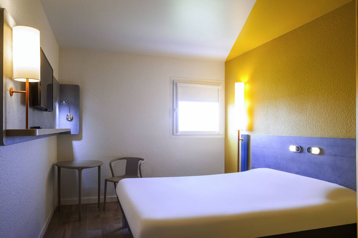 Ein Hotelzimmer mit Bett, kleinem Tisch, Stuhl, Spiegel und Fenster.