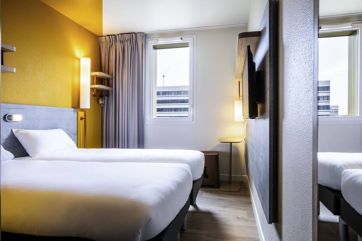 Ein Hotelzimmer mit zwei Betten, einem Fenster mit Vorhängen, einem Nachttisch mit Lampe und einem an der Wand montierten Fernseher.