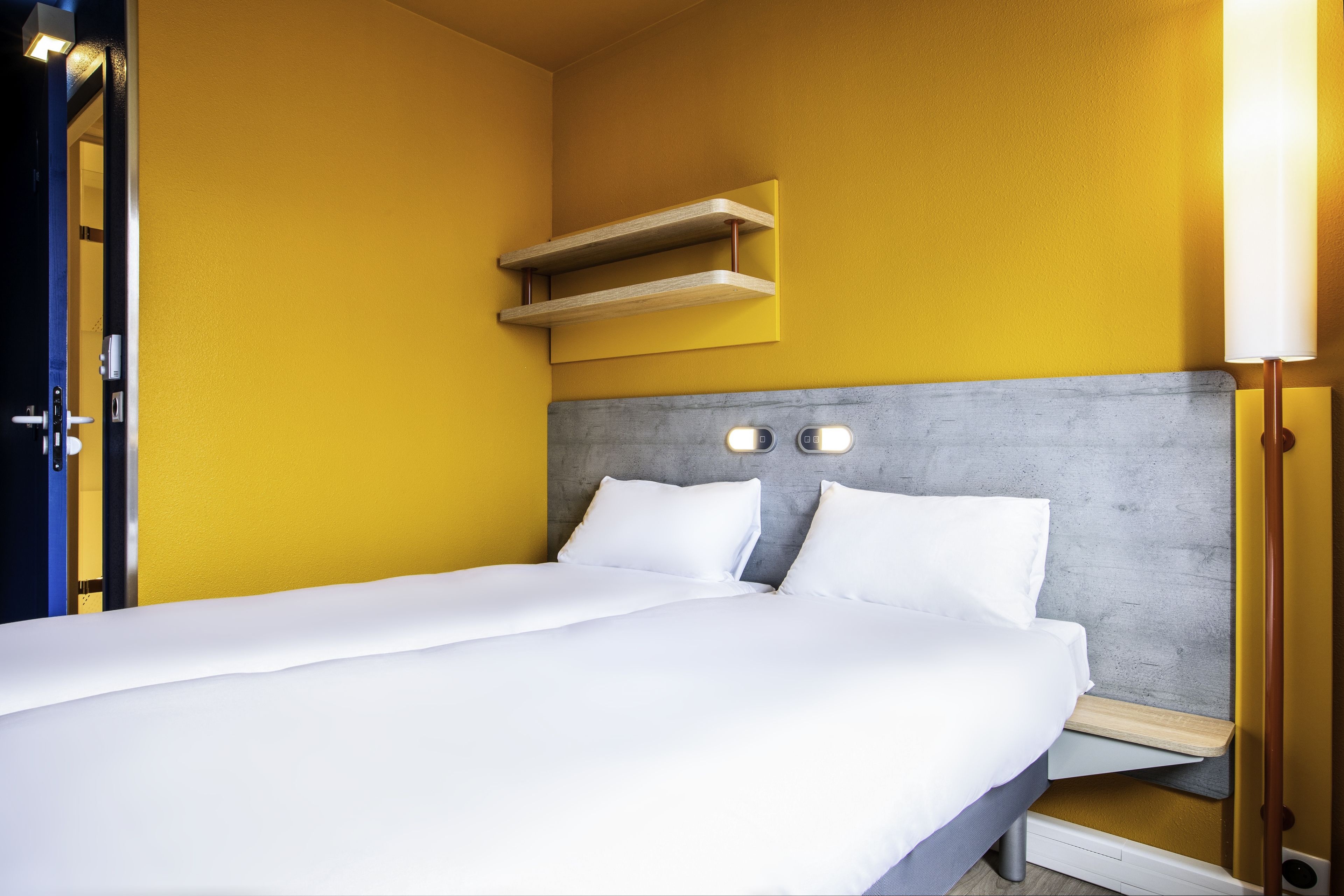 A hotel room with a bed, a bedside table, a shelf above the bed, and a door.