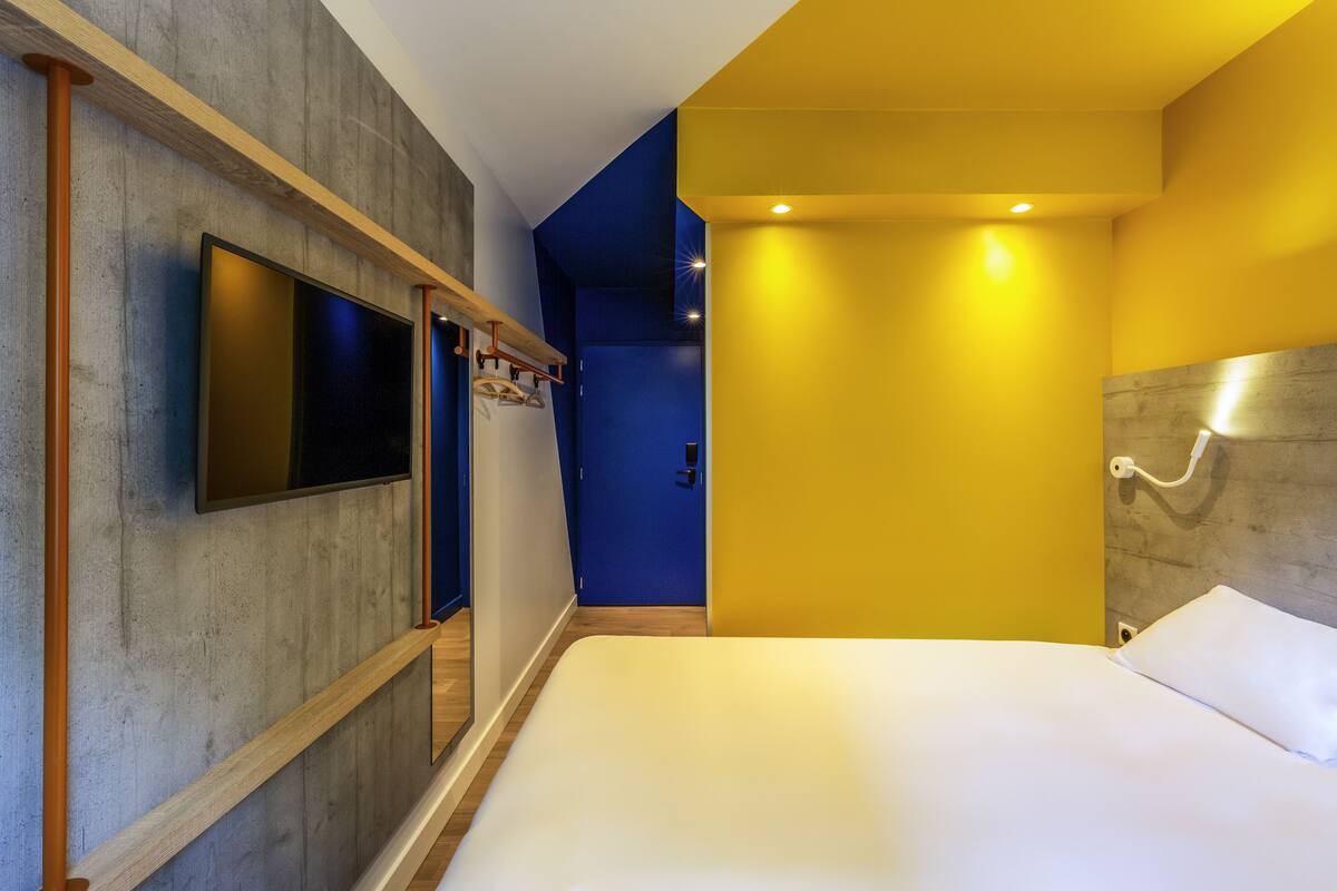 A modern hotel room with a large bed, a flat-screen TV mounted on the wall, and a wooden ceiling beam.