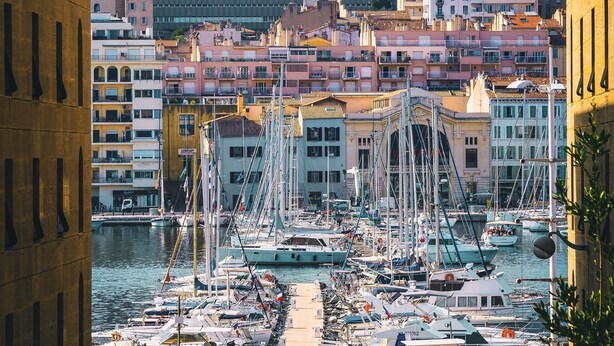 Una marina con barche ormeggiate, edifici sullo sfondo e una collina con case.