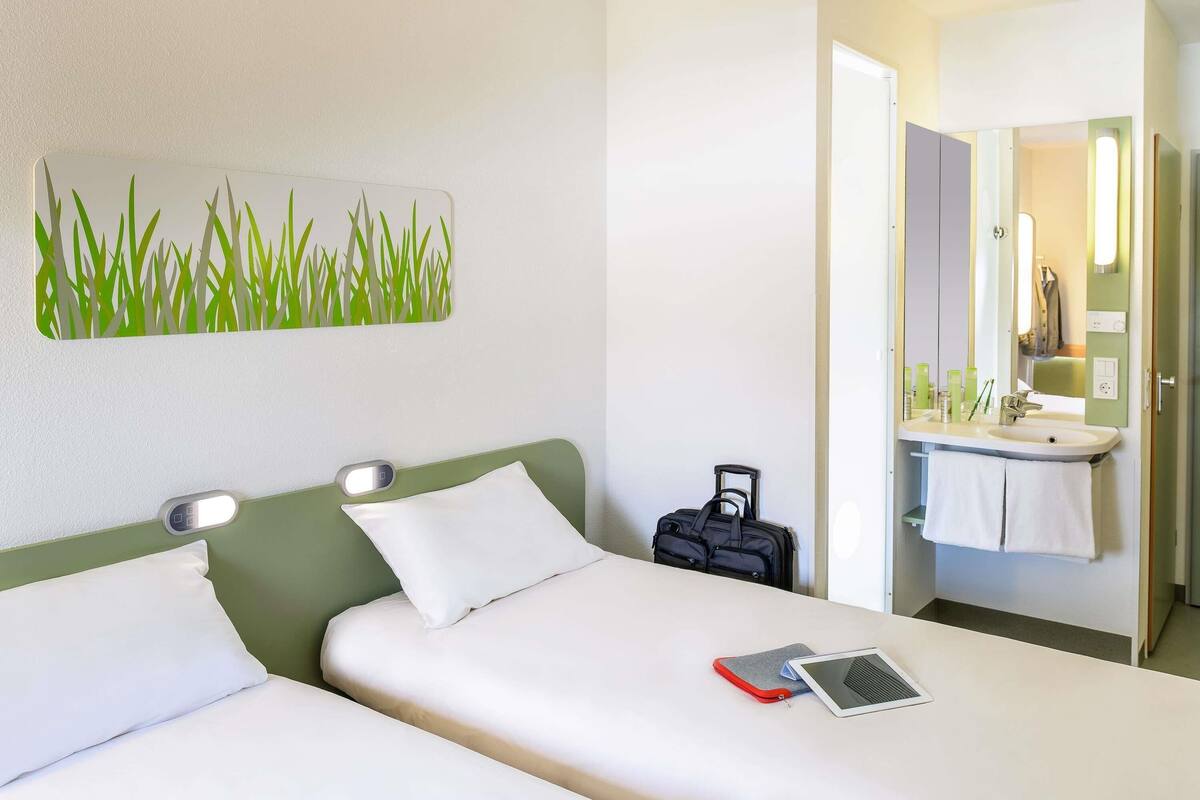 Une chambre d’hôtel comprenant un lit, une table de chevet, une valise, une salle de bain avec un lavabo et un miroir, ainsi qu’une œuvre d’art murale.