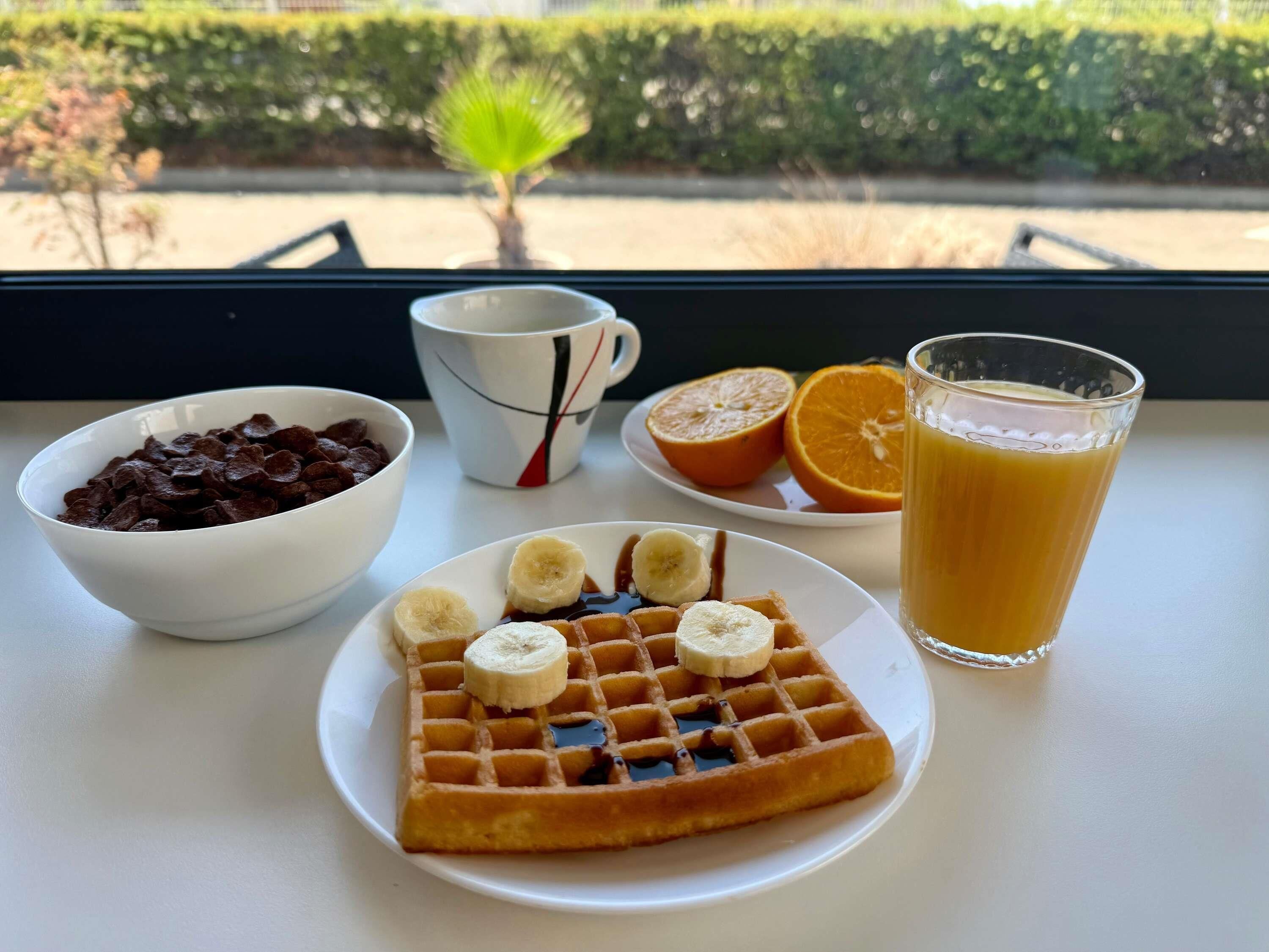 A breakfast setting with waffles, bananas, orange slices, a glass of orange juice, and a bowl of chocolate cereal on a table by a window.