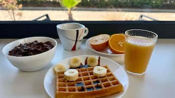 Desayuno con waffles, plátanos, gajos de naranja, un vaso de jugo de naranja y un tazón de cereal de chocolate sobre una mesa junto a una ventana.
