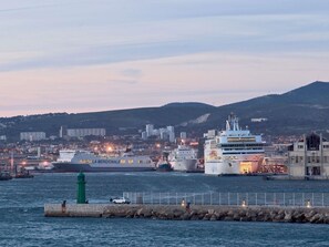 Ein Hafen mit großen Kreuzfahrtschiffen, einem grünen Leuchtturm und einer Stadtkulisse im Hintergrund.
