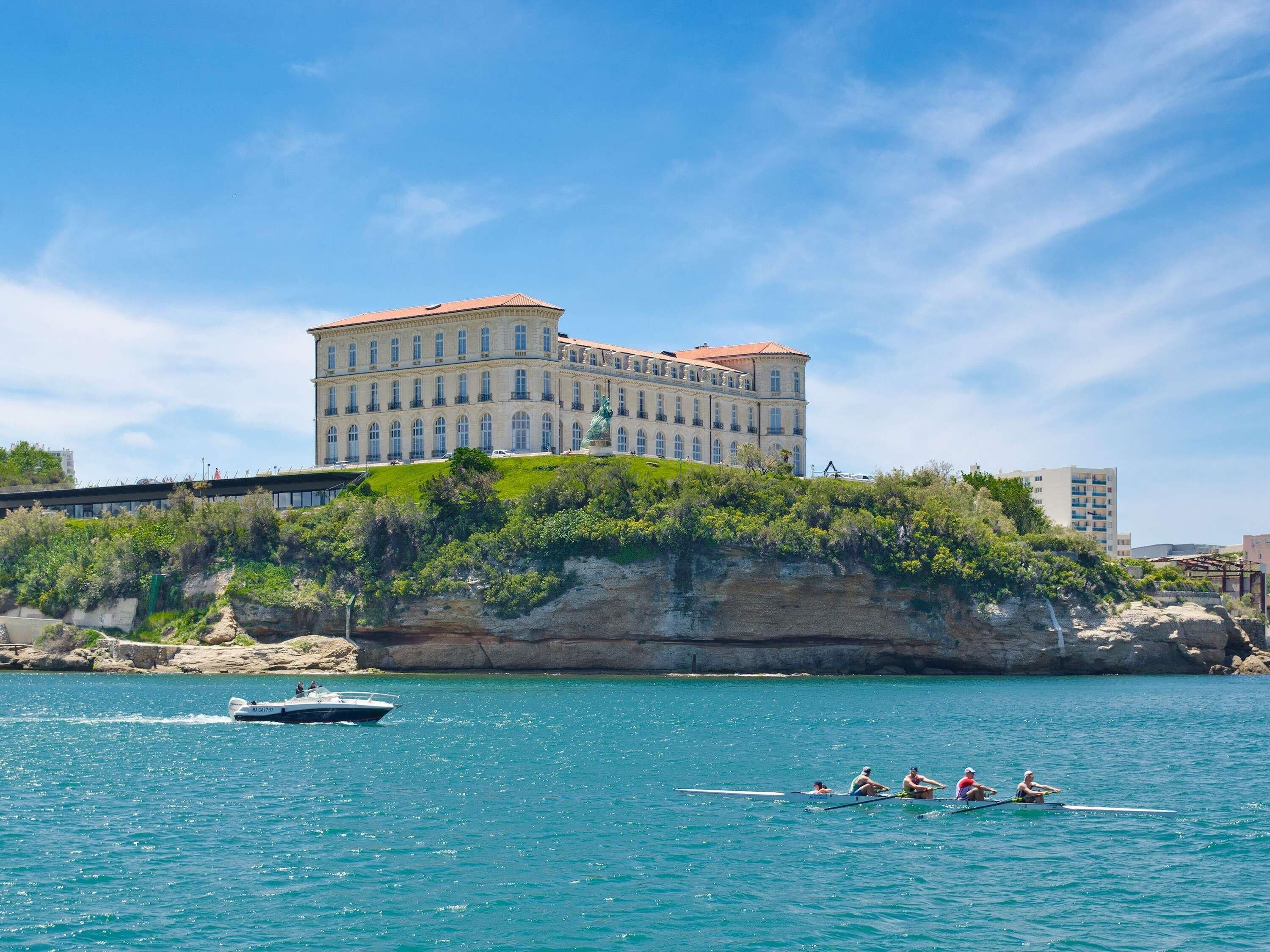 Un gran edificio histórico con un balcón destacado, situado junto a una masa de agua tranquila con barcos y remeros.