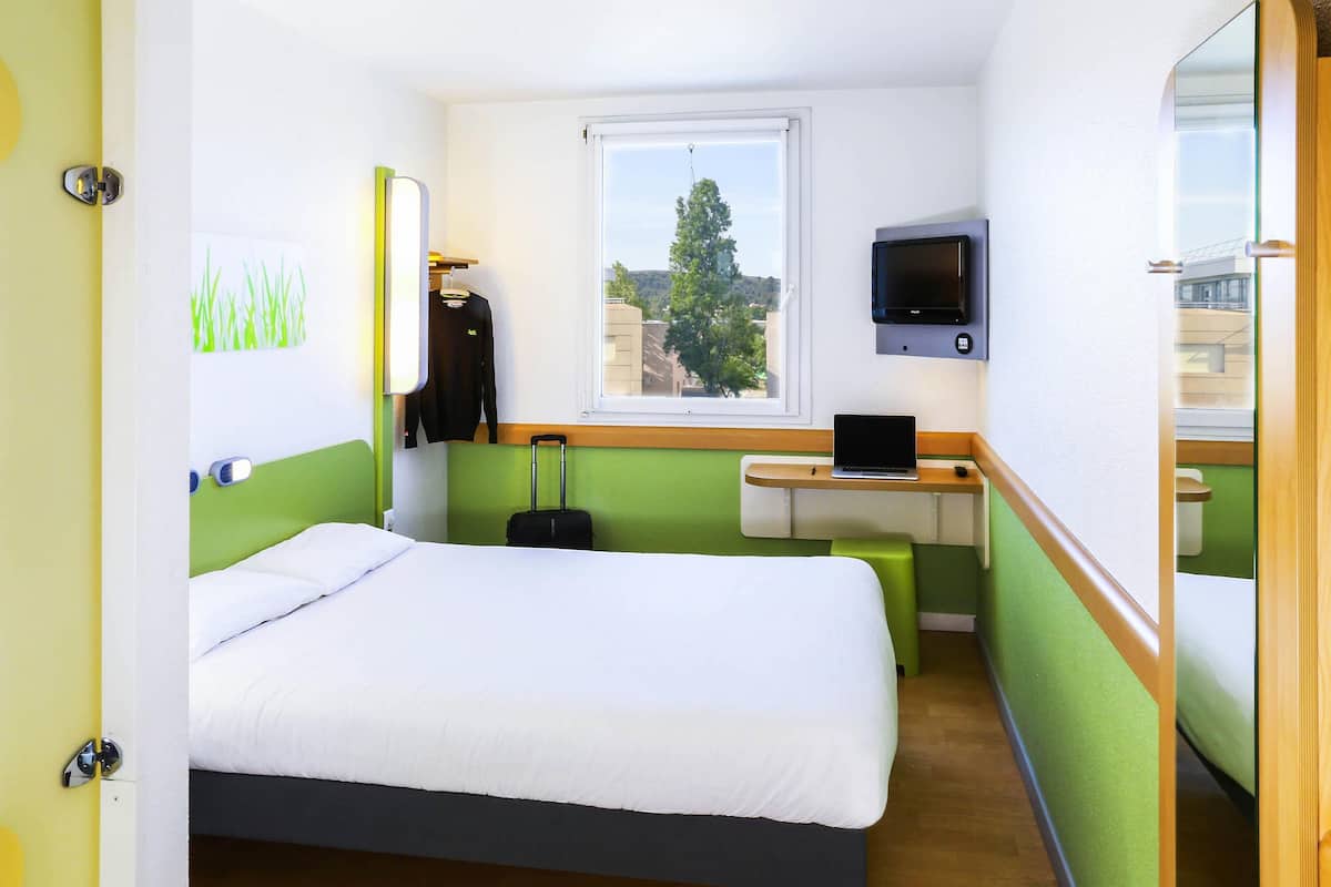 Quarto de hotel com uma cama, uma escrivaninha com um laptop, uma televisão, uma mala e uma janela com vista.