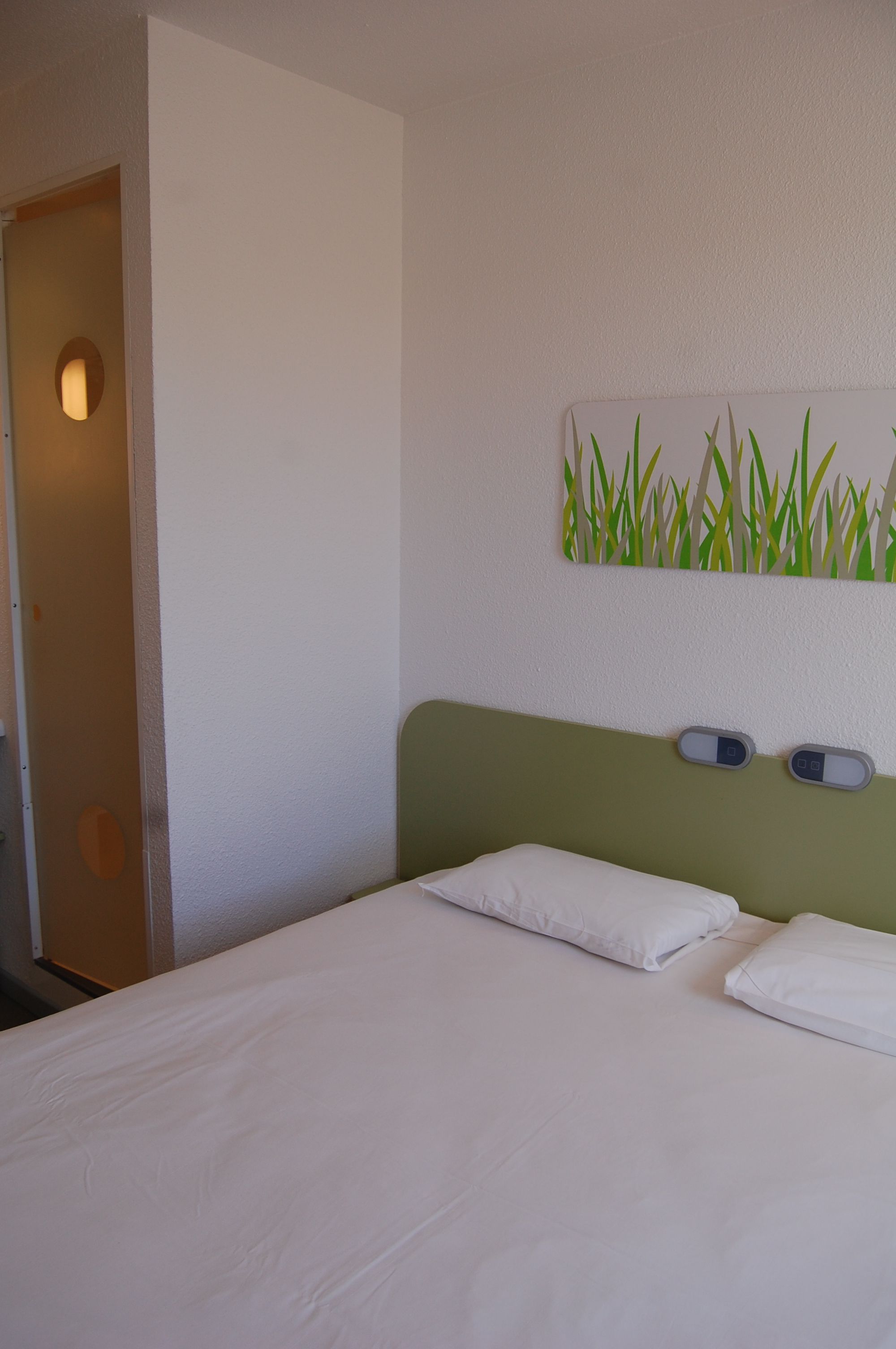 Una habitación de hotel con una cama, cabecera verde y un espejo fijado a la pared.