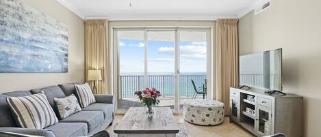 A living room with a sofa, coffee table, and TV stand, a balcony with a view of the sea, and a ceiling fan.