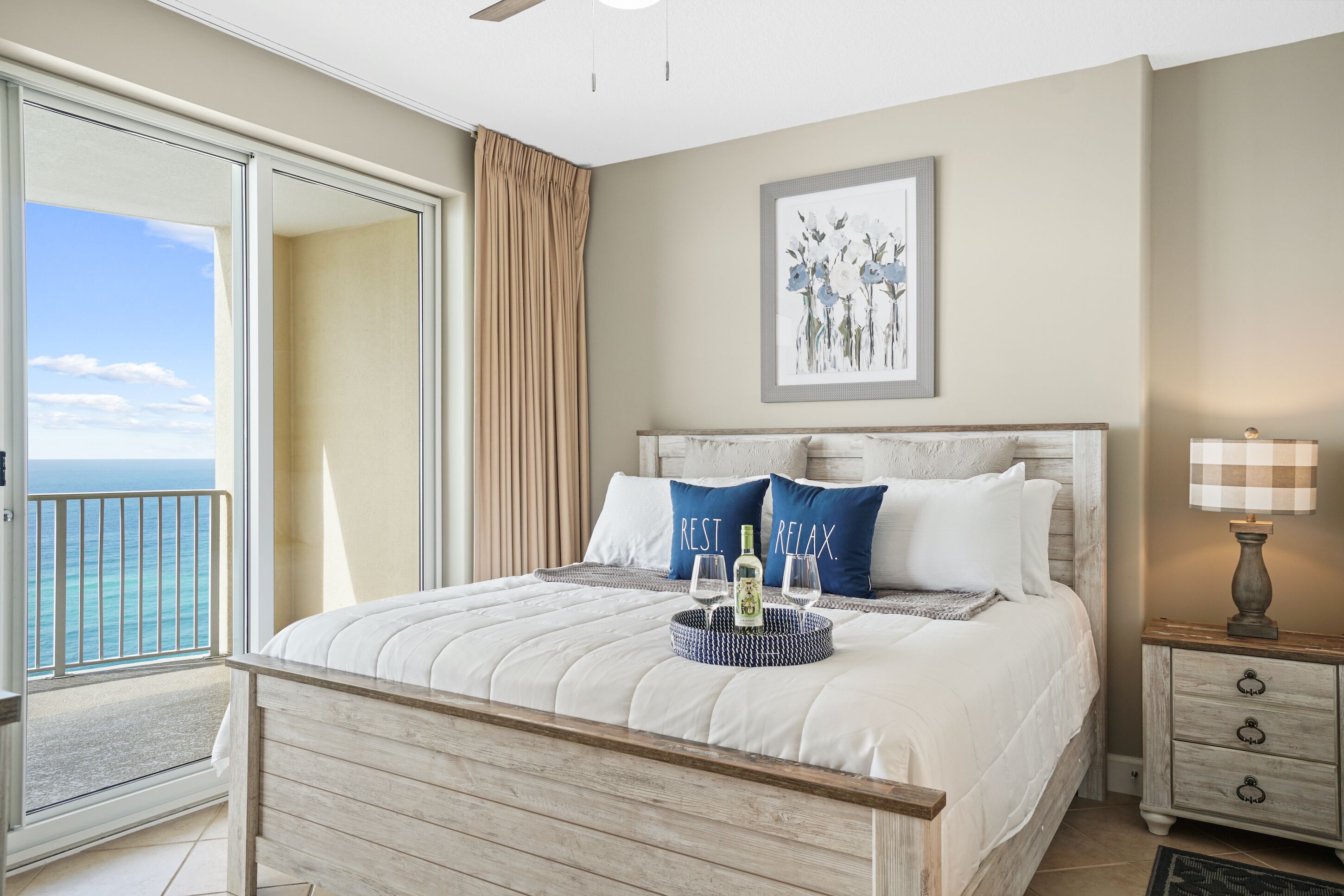 A bedroom with a bed, a nightstand, a lamp, and a view of the ocean.