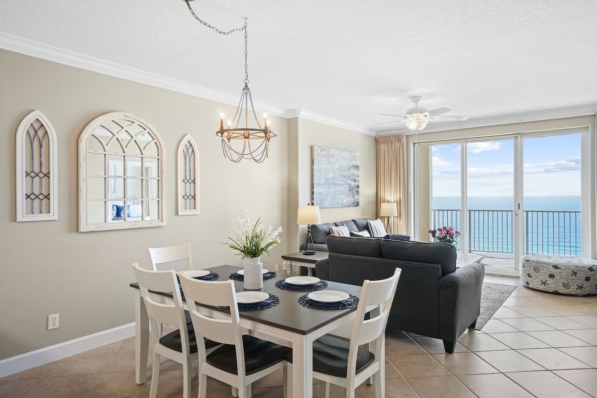 A dining area with a table set for four, a chandelier, and a view of the ocean through large windows.