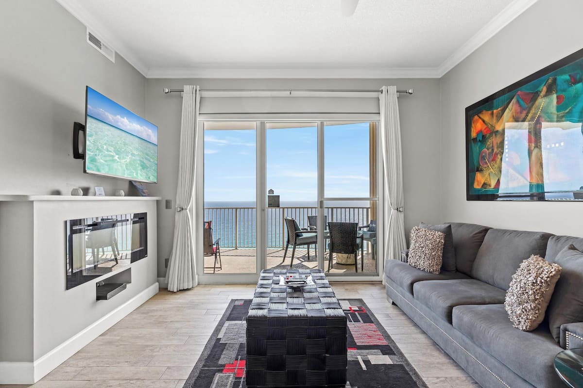 A living room with a TV, a sofa, a coffee table, and a balcony with a view of the sea.