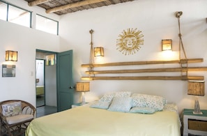 Un dormitorio con cama, dos mesitas de noche con lámparas, una silla de mimbre, un muro con un diseño de sol y una puerta que da a otra habitación.