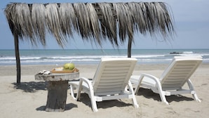 A beach scene with a thatched roof structure, two white lounge chairs, and a small table with a bowl and a green fruit. - La Casa los Tumpis (Zorritos)