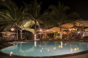 Zona de piscina nocturna con palmeras, sillones de descanso y sombrillas.