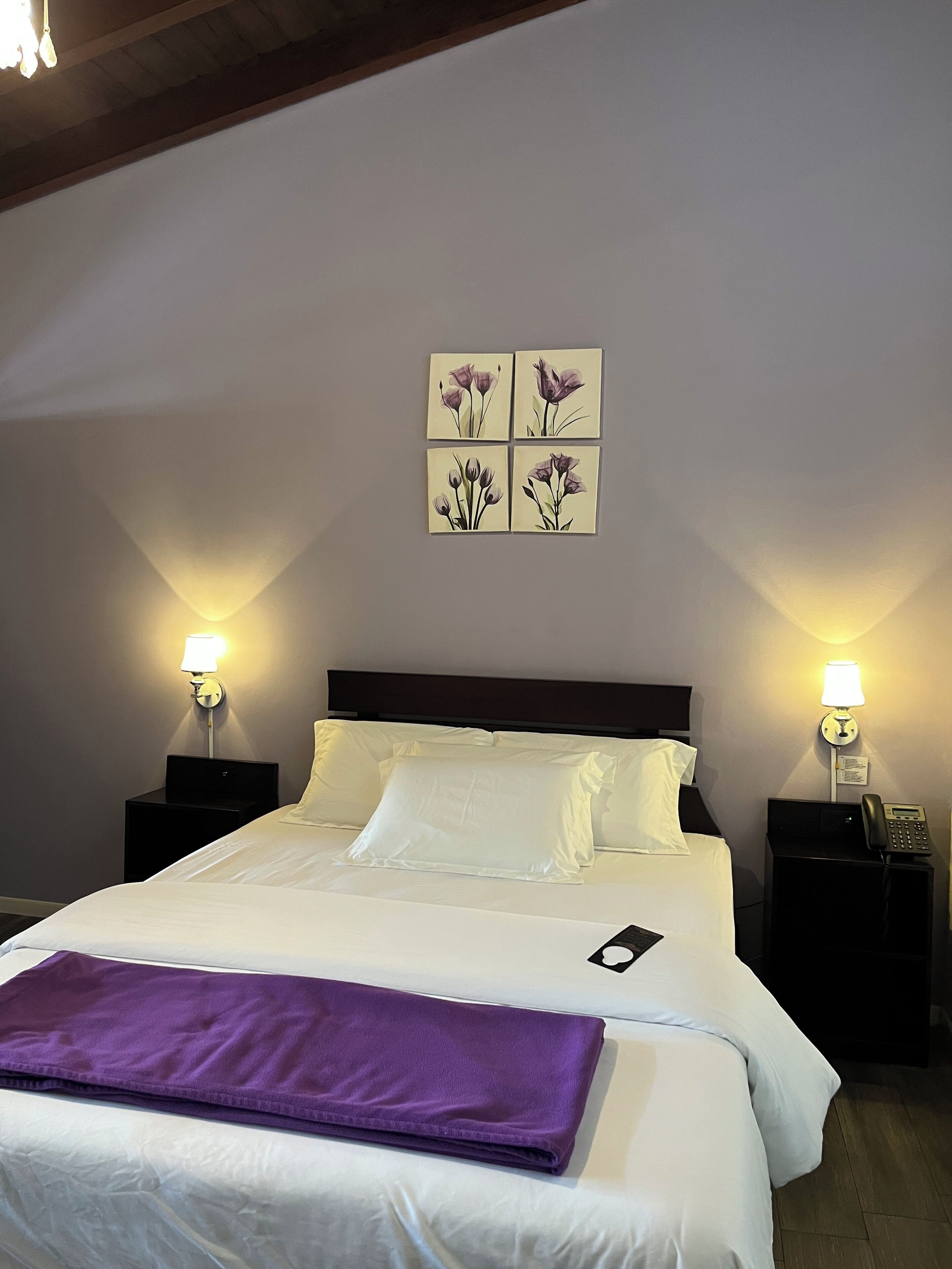 Habitación de hotel con cama, dos mesitas de noche con lámparas, teléfono en la pared y una manta púrpura sobre la cama.