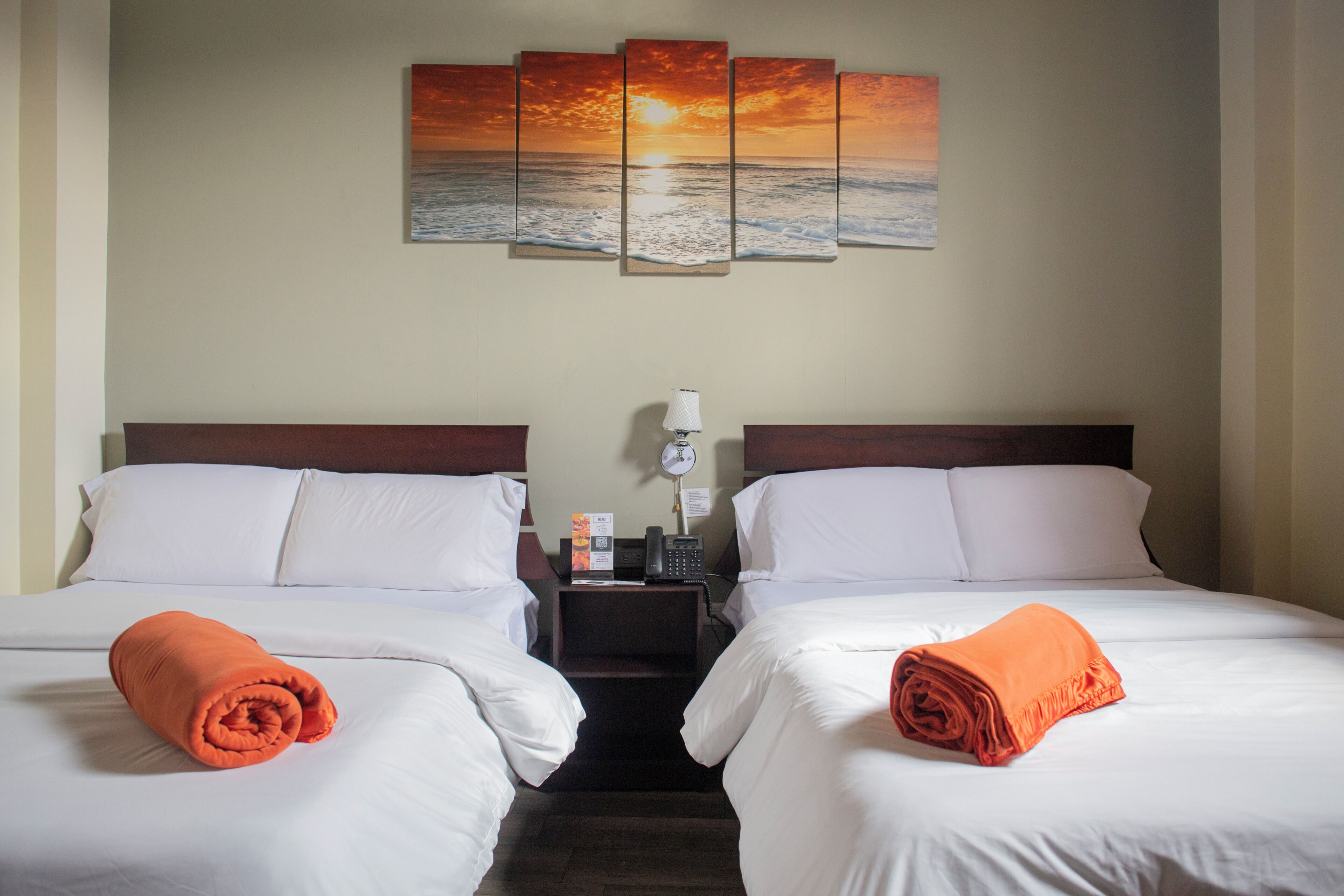 Habitación de hotel con dos camas, una mesita de noche con lámpara, teléfono y un cuadro con atardecer sobre el océano.