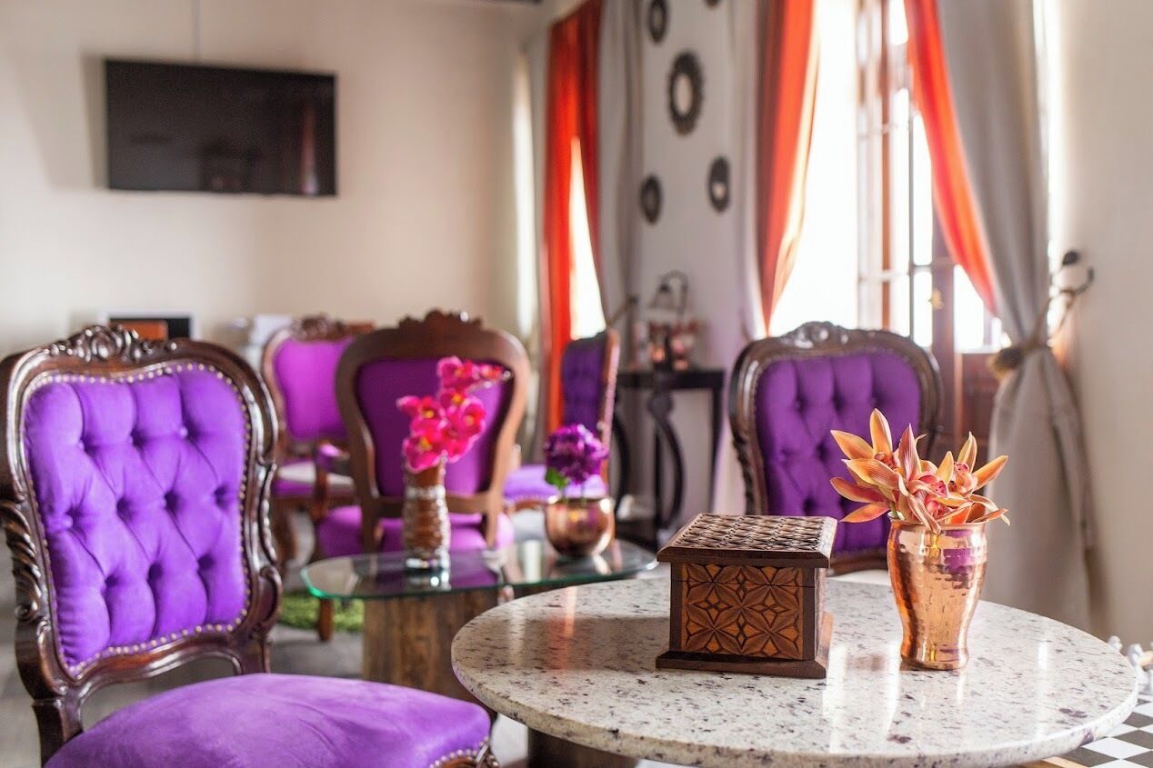 Una habitación con sillas tapizadas de color morado, una mesa redonda con un estuche decorativo y un florero con flores, y vistas al exterior a través de amplias ventanas con cortinas naranjas.