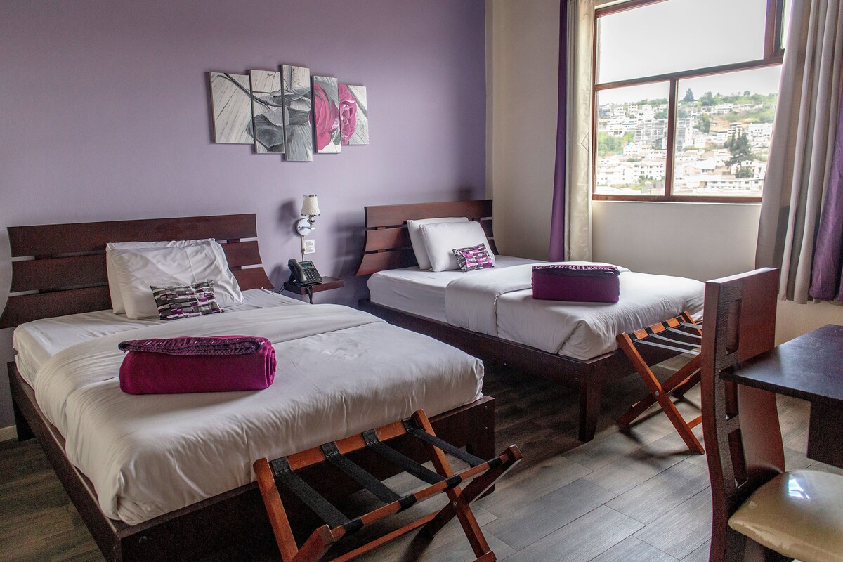 Habitación de hotel con dos camas, una silla de madera, una mesa y vistas a un paisaje urbano a través de una ventana.