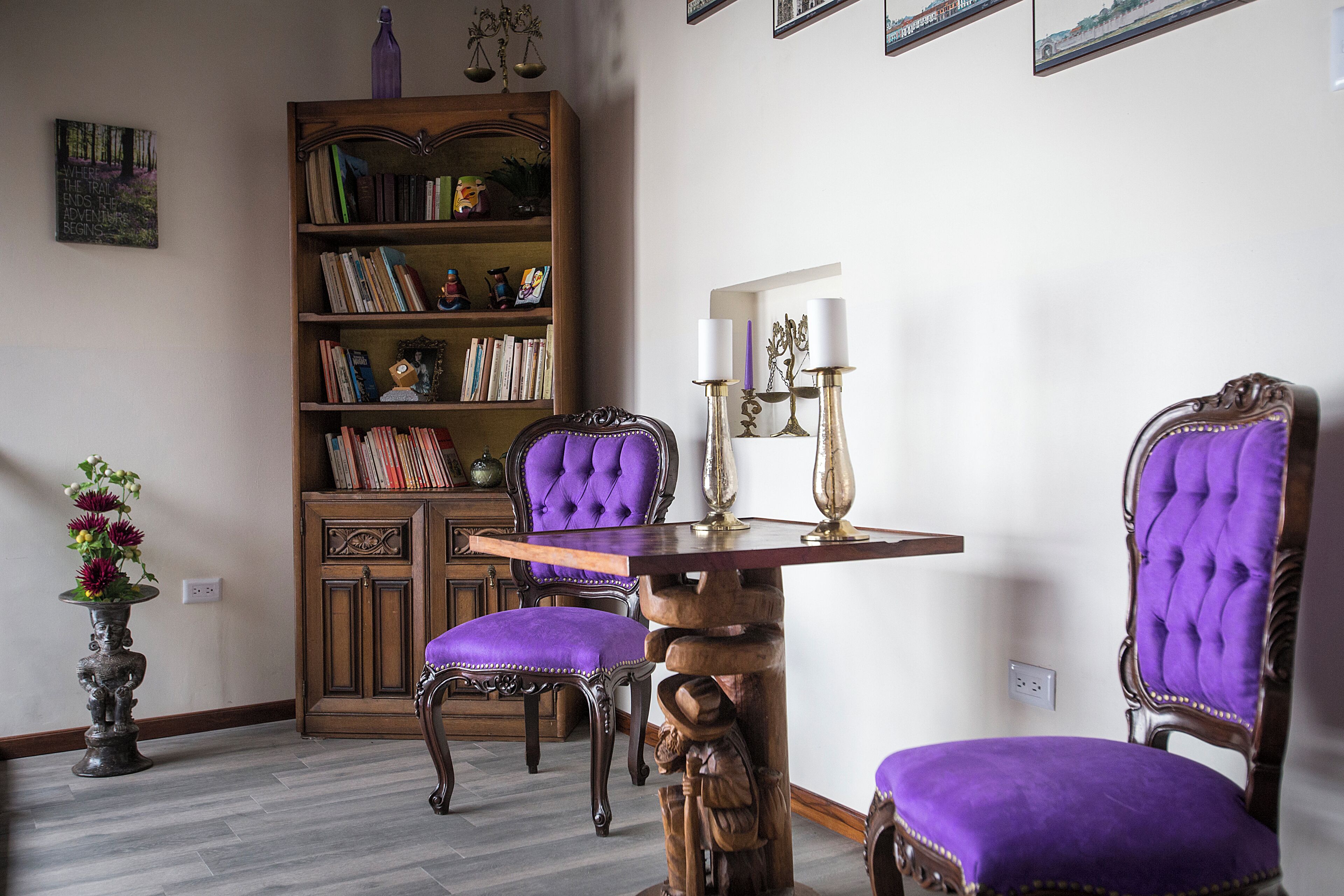 Una habitación con una estantería de madera, dos sillones tapizados en morado, una mesita con velas y un jarrón decorativo.
