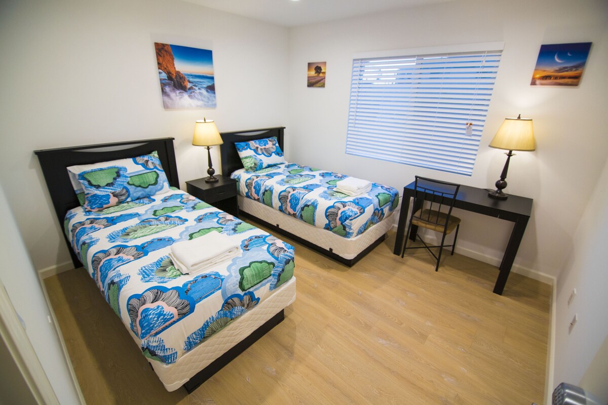 A hotel room with two beds, each with a matching headboard and bedside table, a desk, and a window with blinds.