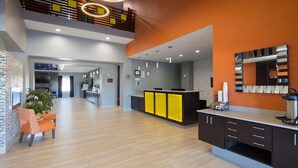 Um saguão moderno de hotel com balcão de recepção, área de estar com cadeiras laranja e uma cozinha ao fundo.
