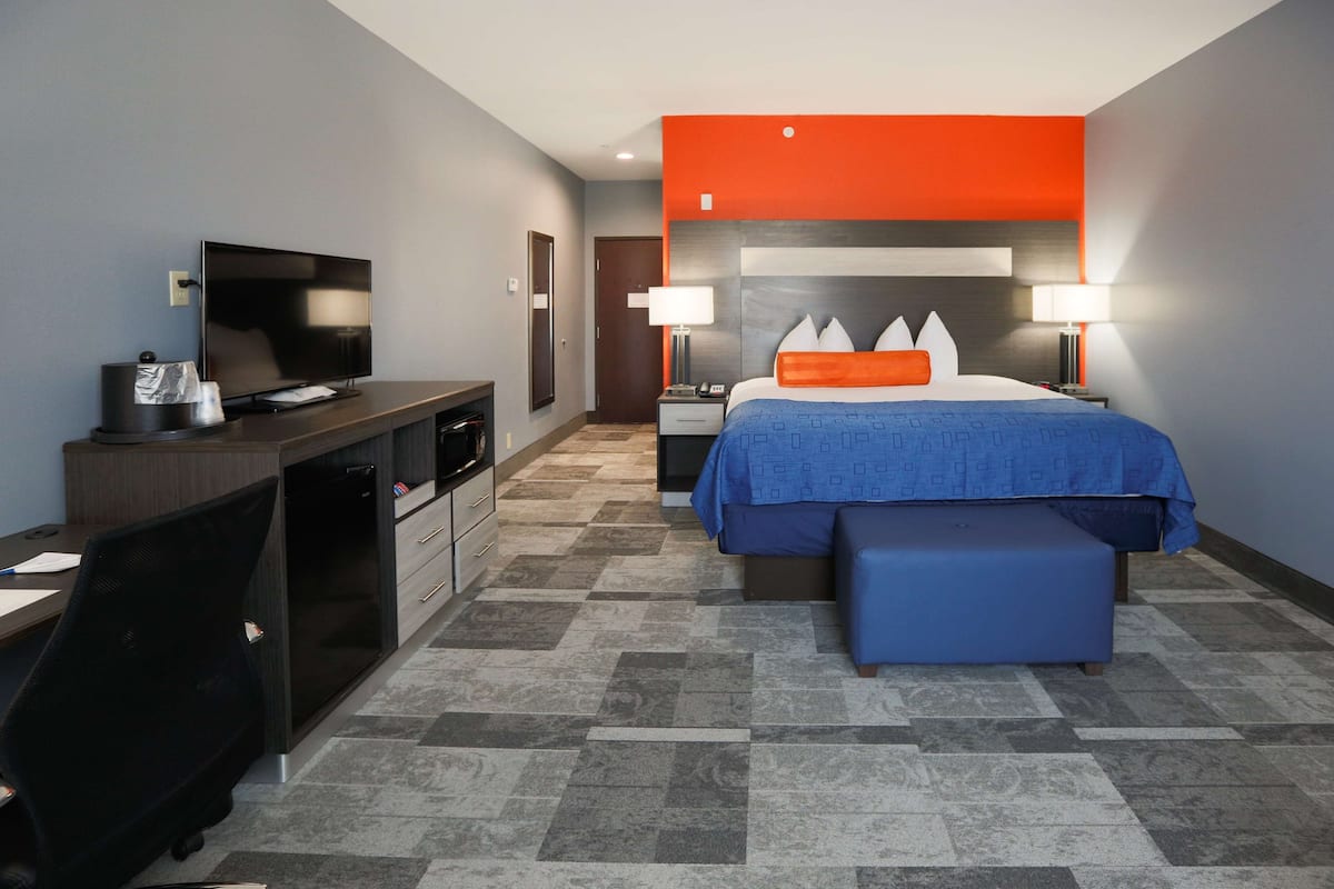 A hotel room with a bed, a desk with a TV, a chair, and a small table with a lamp.