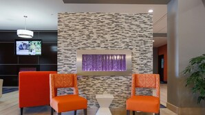 Um saguão com duas poltronas laranja, uma televisão na parede e um painel decorativo com luz roxa.