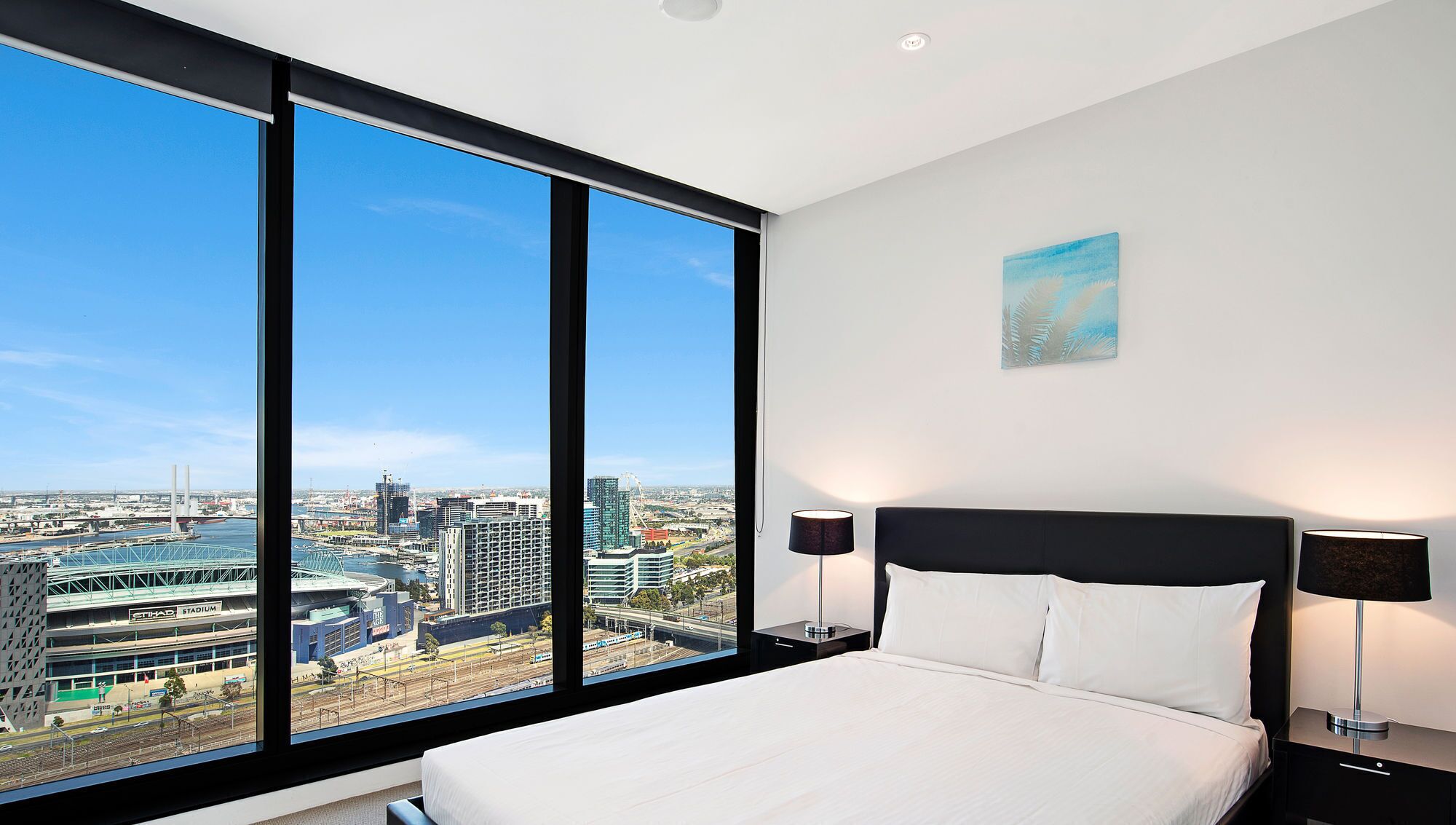 A hotel room with a large window, a bed, a nightstand, a lamp, and a painting.