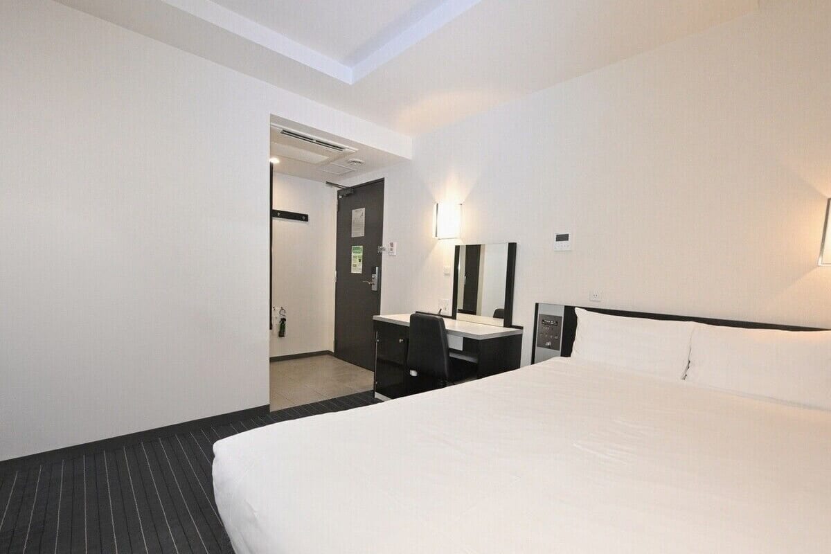 A hotel room with a large bed, a desk with a chair, a mirror, and a door leading to another room.