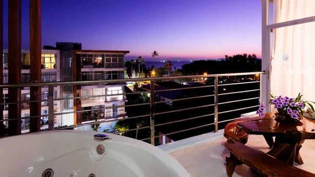 Un balcón con un jacuzzi, una mesa de madera con flores y vistas a edificios y al atardecer.