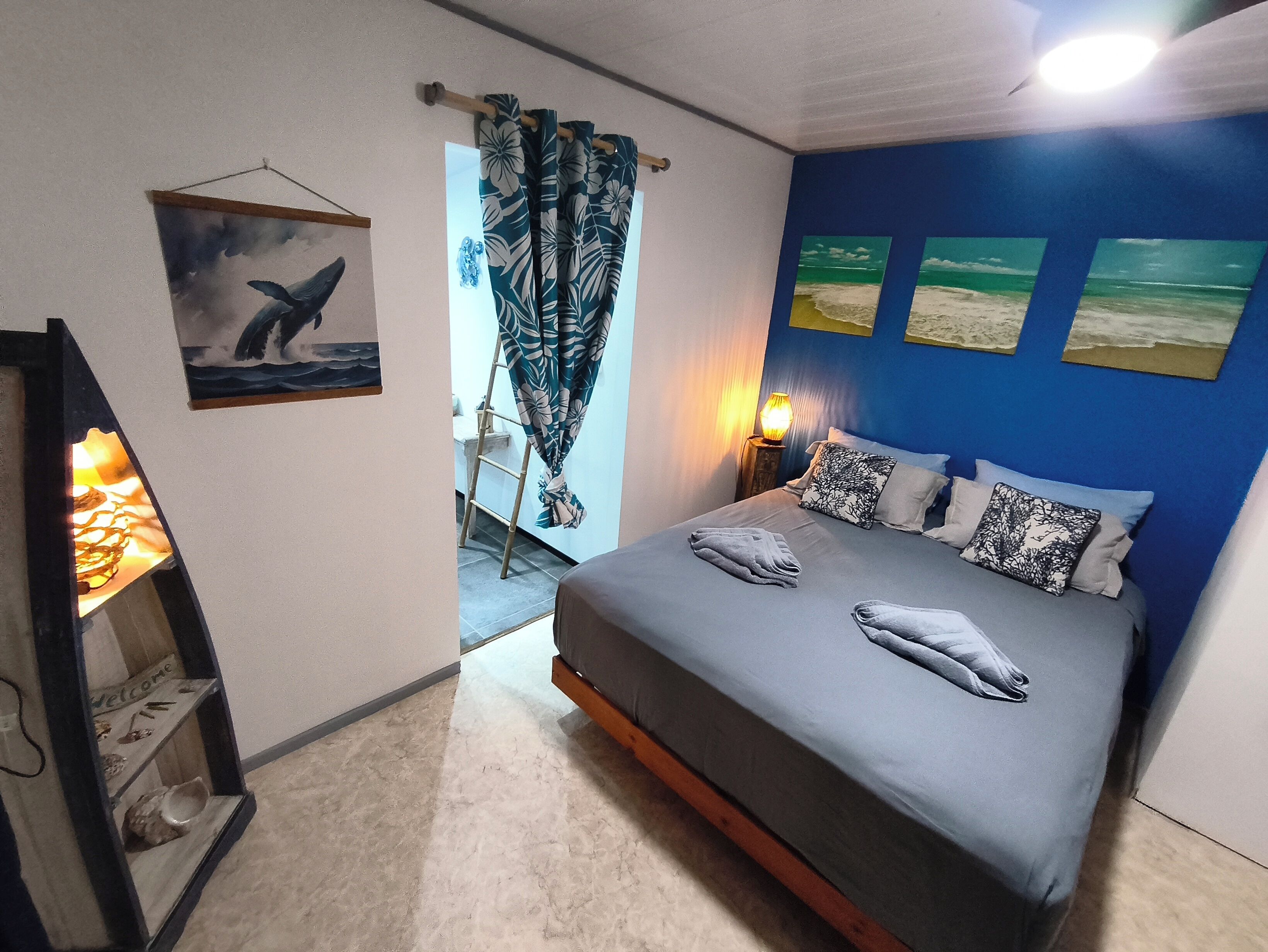 Un dormitorio con cama, almohadas, mesita de noche, lámpara de pared y un cuadro enmarcado.