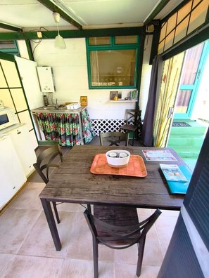 A dining area with a wooden table, chairs, a small kitchen area with a microwave, and a window with curtains. - Te Ava Piti Lodge (Raiatea)