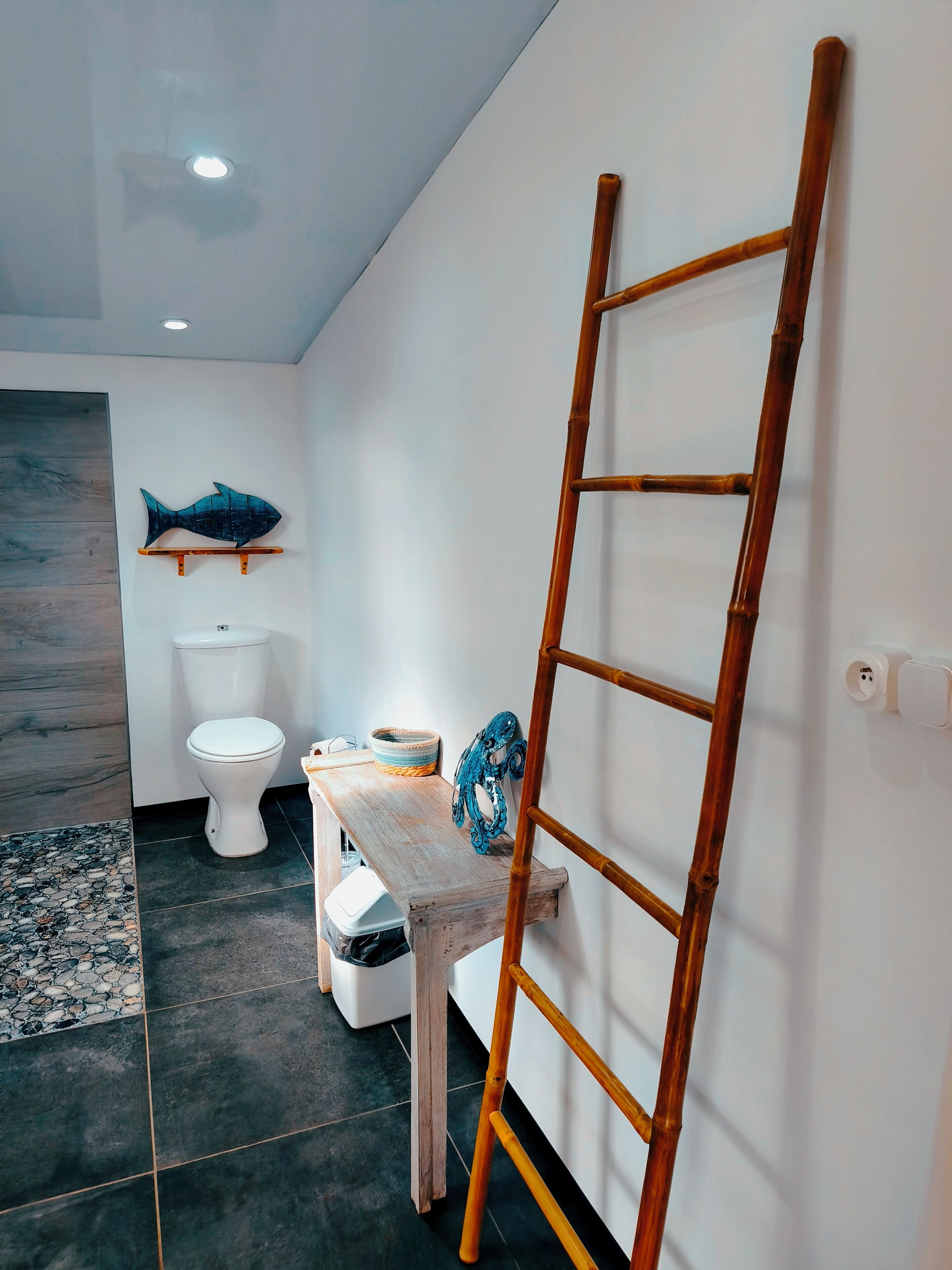 Ein Badezimmer mit Toilette, einer Holzbank, einer Leiter, einem an der Wand befestigten Regal mit Fischverziierung und einem Fliesenboden.