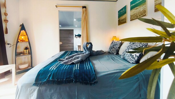 A neatly made bed with a blue blanket and pillows, a small shelf with a lamp, and a view of the room's interior. - Te Ava Piti Lodge (Raiatea)