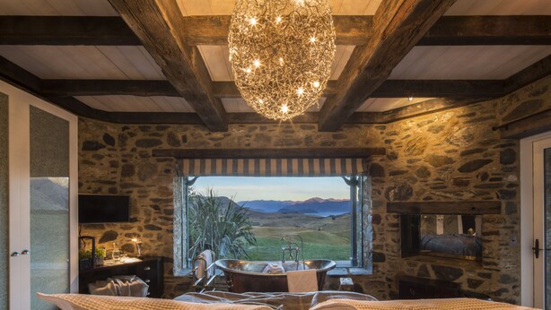 A rustic room with stone walls, a large window with a view of the countryside, a wooden ceiling, and a chandelier. - Mahu Whenua (Treble Cone)