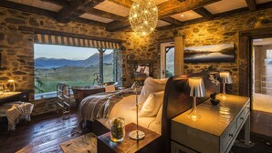A rustic bedroom with a stone wall, wooden beams, a large window with a mountain view, a bed with a white comforter, and a desk with lamps. - Mahu Whenua (Treble Cone)