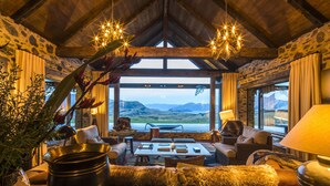 A cozy interior with a stone wall, wooden ceiling, and a large window offering a scenic view. Furnishings include a sofa, chairs, and a table with a candle. - Mahu Whenua (Treble Cone)