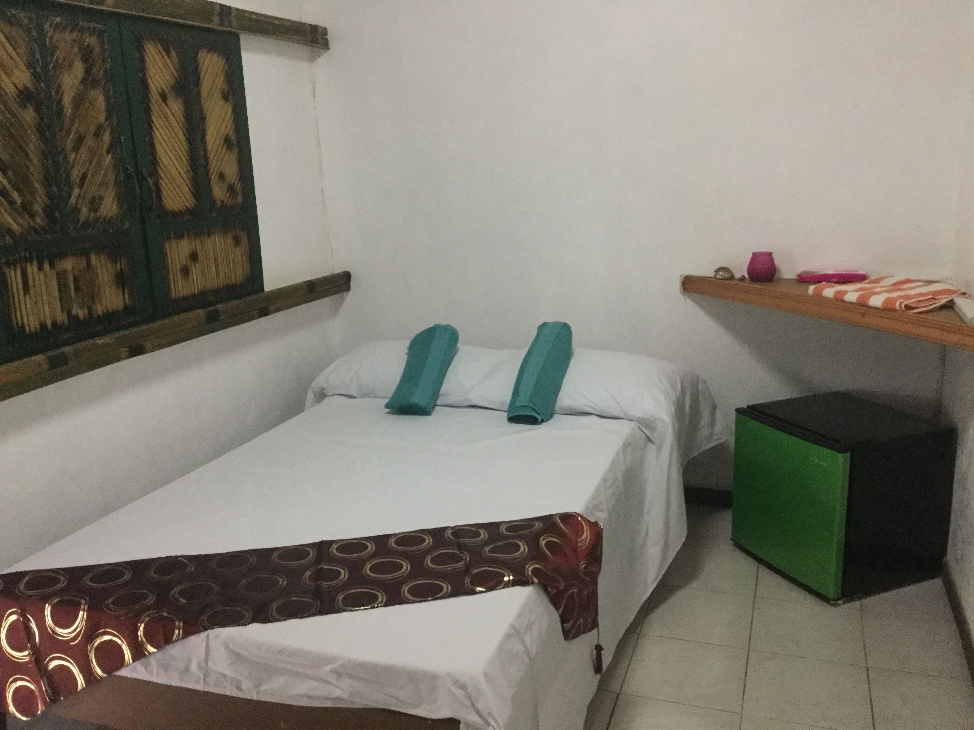 Una habitación pequeña con cama individual, cabecera de madera, un cesto de basura verde y una estantería con objetos personales.