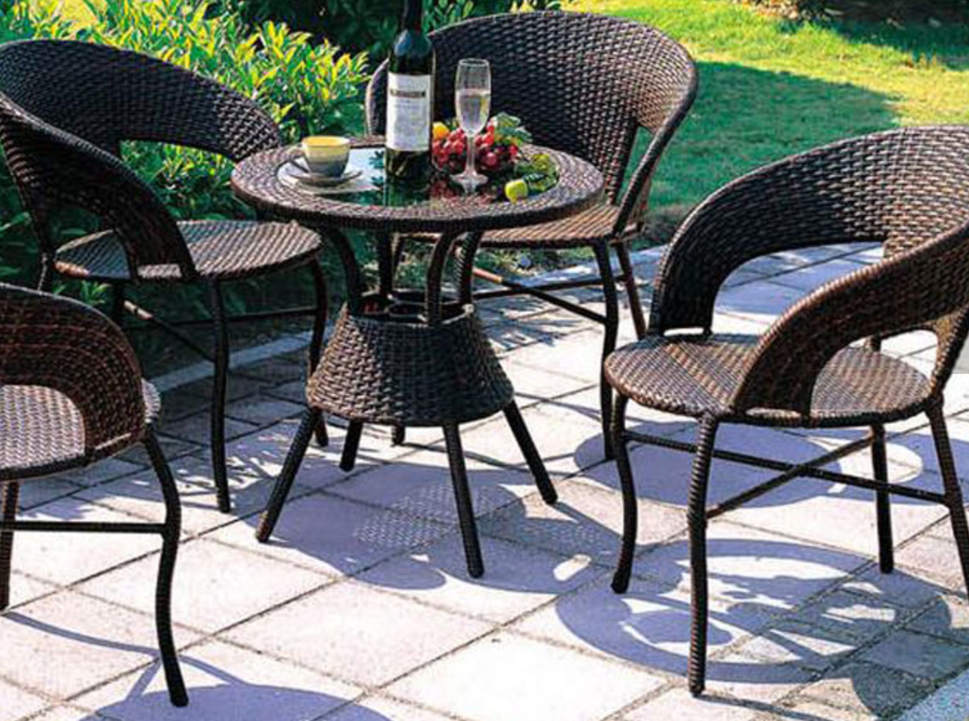 An outdoor patio with wicker chairs and a round table, featuring a bottle, glass, and fruits.