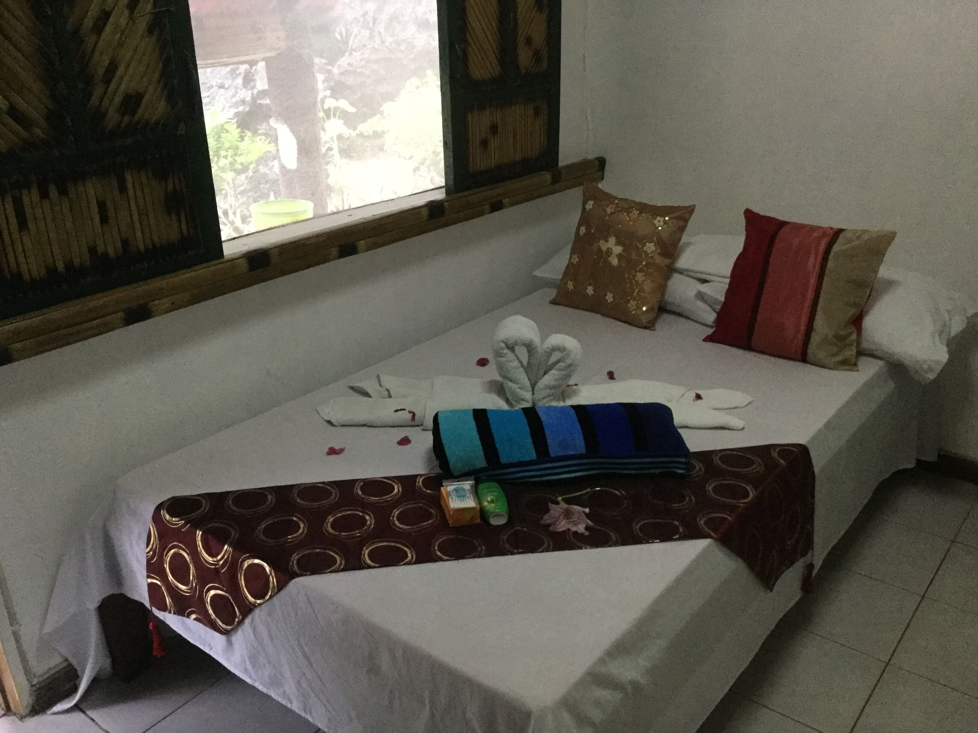 A bed with a white sheet, a brown patterned blanket, and a blue striped pillow. There are two pillows on the bed, one with a floral design and another with a red and beige pattern. A small green bottle and a pink flower are on the bed.