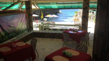 Un restaurant doté d’une terrasse offrant une vue sur la mer, avec des tables recouvertes de nappes rouges et des chaises à motifs.