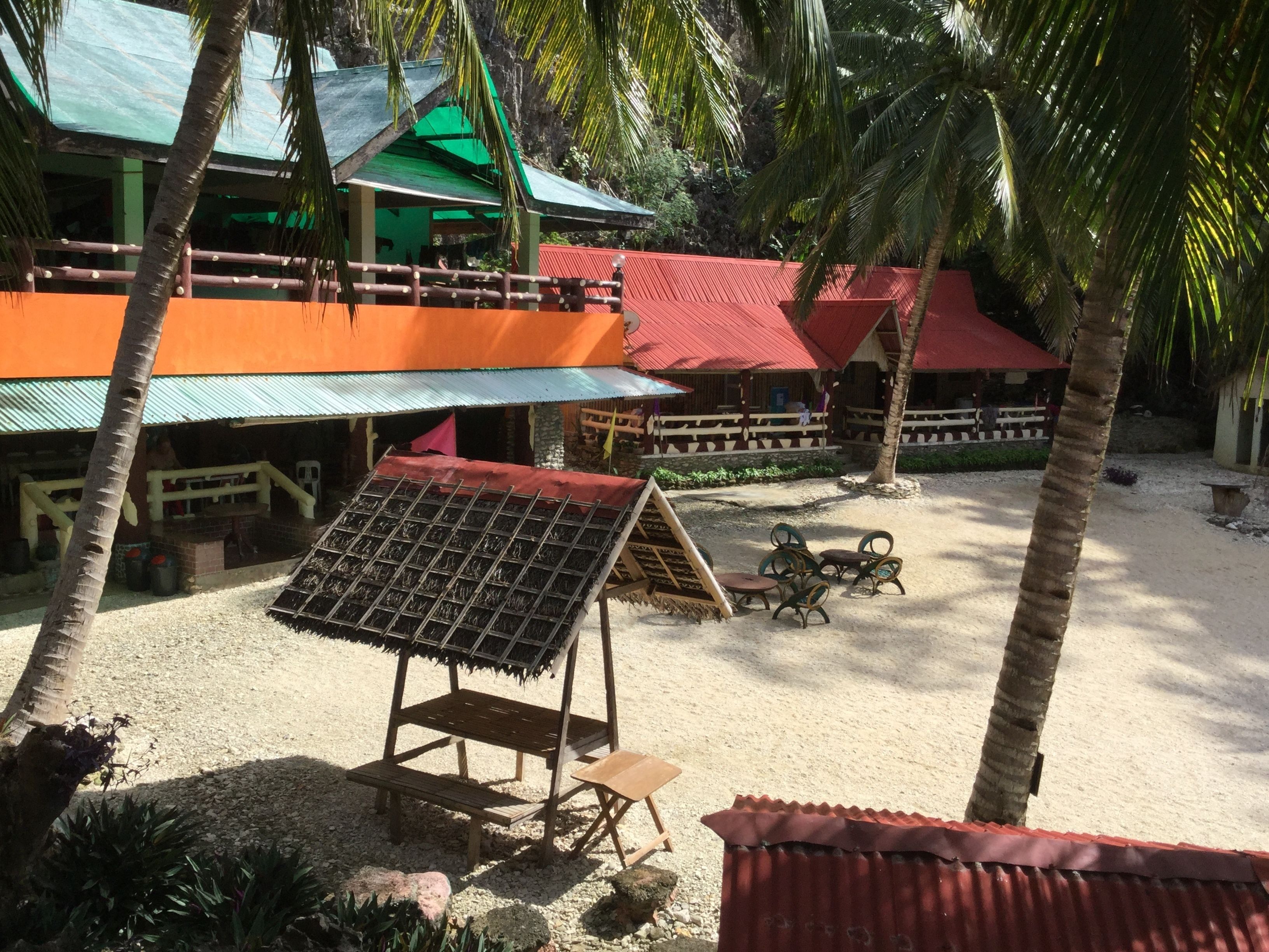A beachside resort with multiple buildings, palm trees, and outdoor seating areas.