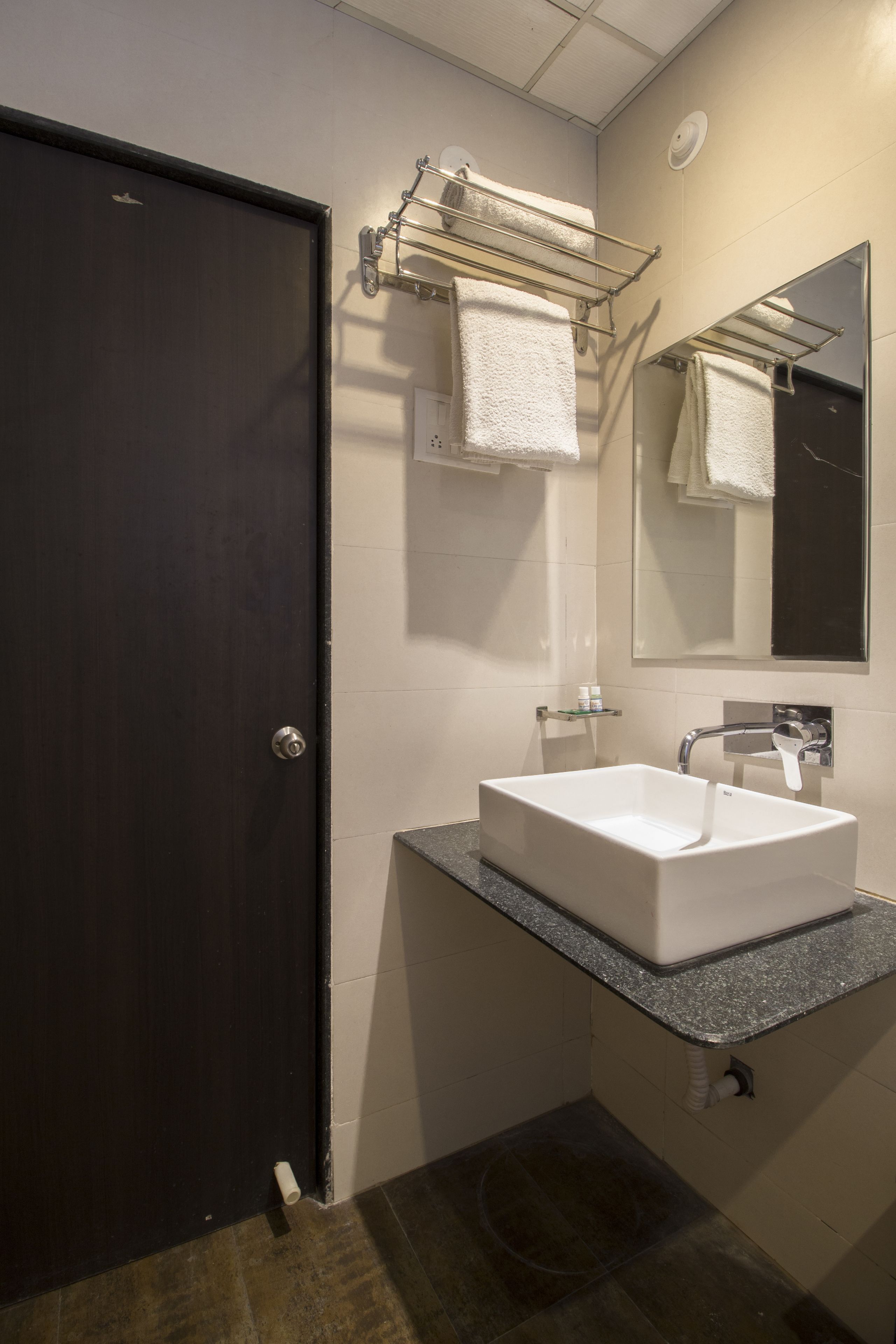 A bathroom with a dark door, a white sink, a towel rack, a mirror, and a towel dispenser.
